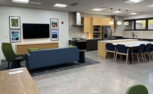 Image of the family room space of the learning lab that includes a blue coach and green chair facing a wall with a large screen tv adhered to it and 4 pictures framing it. The kitchen space of the learning lab is to the right and features a large island with 4 chairs around it and various cabinets with a refrigerator, two microwaves and an oven and range lining the wall.