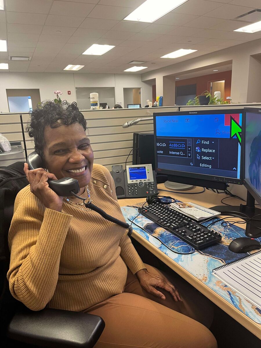 Joyce holds up the desk telephone to her ear while facing the camera and smiling. Behind her are her desktop computer screens. 