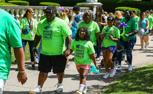 A man holds a young girl's hand as they walk with a crowd on the white cane walk pathway. The little girl has a big smile on her face! Most of the crowd has bright green t-shirts on.