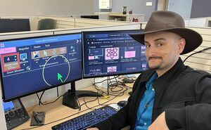 A man wearing a wide brim hat at a desk has assistive technology software pulled up on his computer screen. The curser is large and bright green, with a yellow circle surrounding it, and the screen shows a zoomed in settings page. 