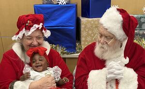 Blind Santa and Mrs. Claus sit together as Mrs. Claus holds a baby dressed up for her photo with Santa. The baby is smiling and wearing a big red bow and red tutu.