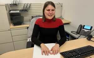 A woman sits in her office and uses both hands to read braille embossed on white paper. 