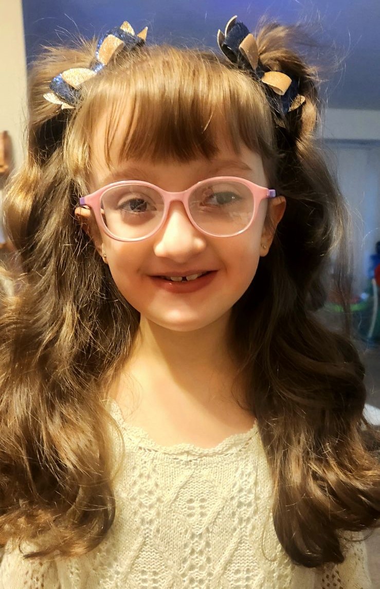 Julianna looks at the camera and smiles. Her long brown hair is in pigtail bows and has pink glasses.