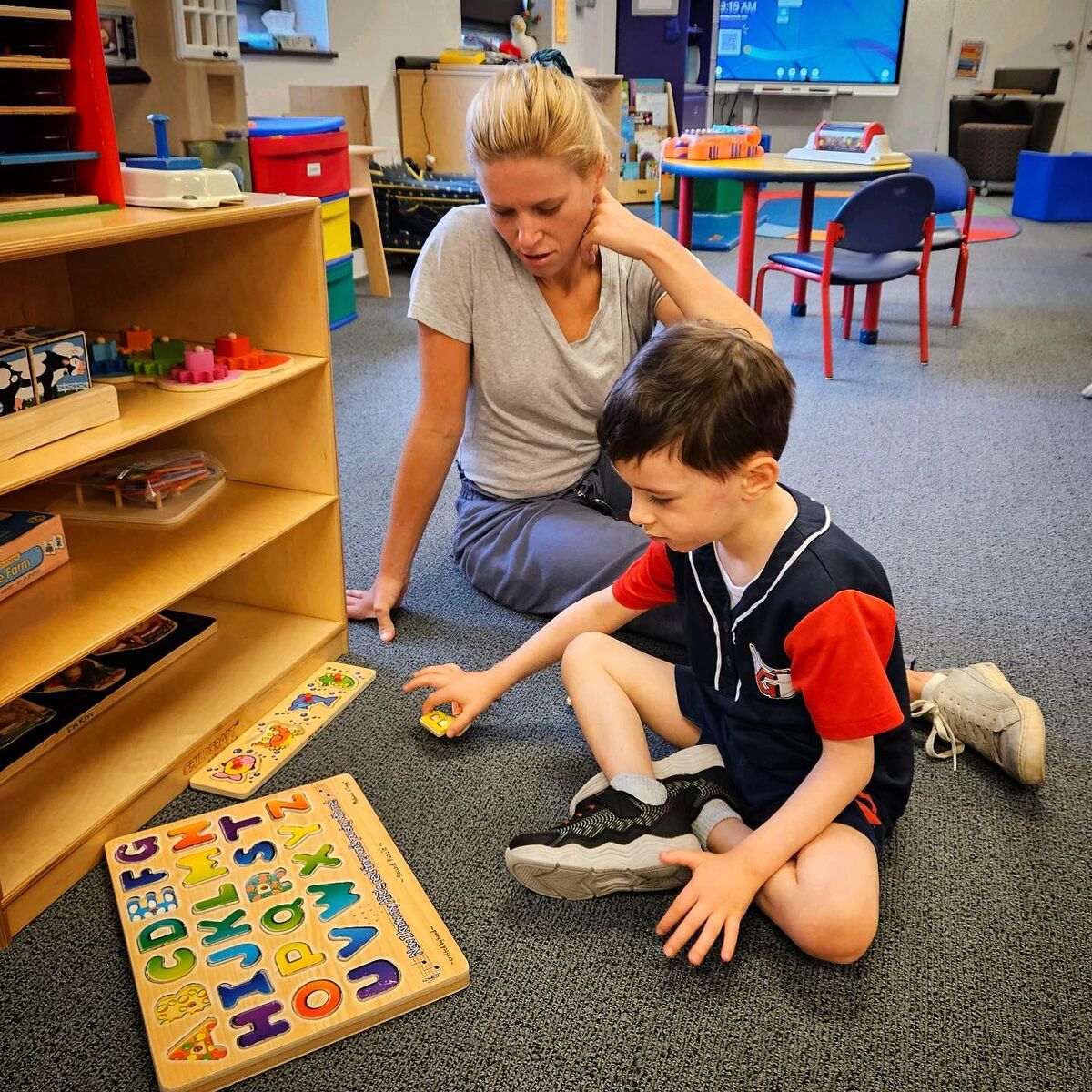 A preschooler plays with a puzzle as a teacher's aid supervises.