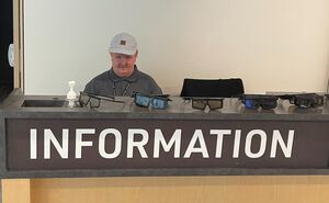 A man sits behind an information desk, wearing a polo shirt and baseball cap.